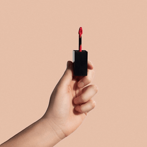 Hand holding premium red MIANIMED skincare lip gloss against a beige background.