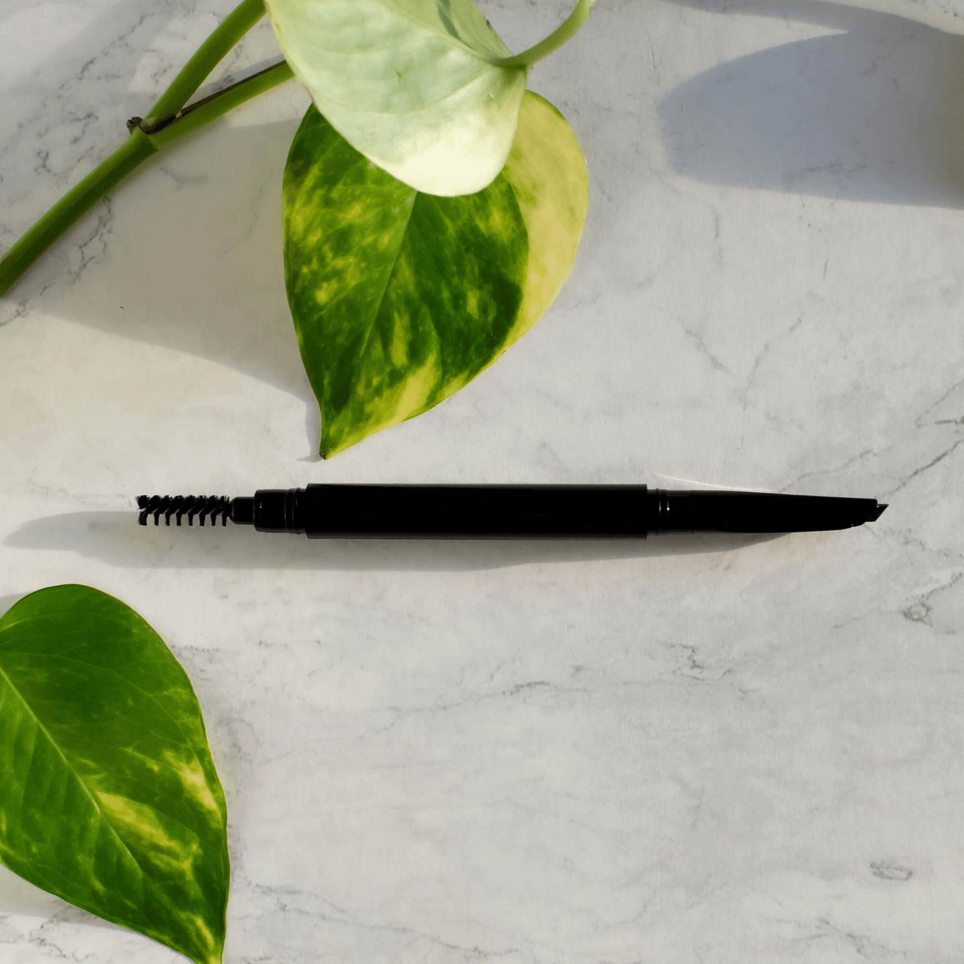 Black eyebrow pencil on marble surface with green leaves, representing premium MIANIMED skincare products.